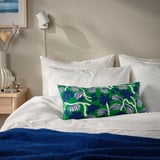 Bed with SANDMOTT green/blue botanical cushion, white linens, blue blanket, wall light, and vase of flowers.
