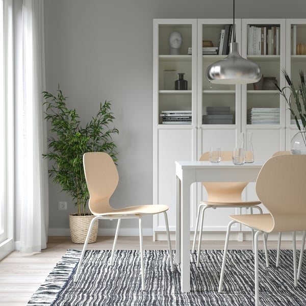 Modern dining area with wooden SIGTRYGG chair, white table, and bookcase. Includes decorative items, minimalist glassware, and a potted plant.