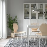 Modern dining area with wooden SIGTRYGG chair, white table, and bookcase. Includes decorative items, minimalist glassware, and a potted plant.