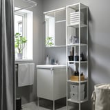 Modern bathroom: white cabinet, mirror with lights, tall shelving with towels, window with plant, gray walls, shower curtain.