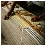 Two people are hand-weaving a wool rug on a loom, creating a pattern with shades of grey and yellow.