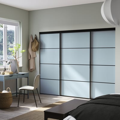 This image a modern, light gray sliding closet, SKYTTA, with black frames, mounted on a wall in a room with a desk and chair.