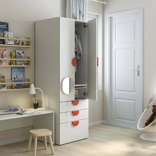 A childs room with a white SMÅSTAD wardrobe, shelves, and desk. The wardrobe has red handles and an open door revealing clothes.