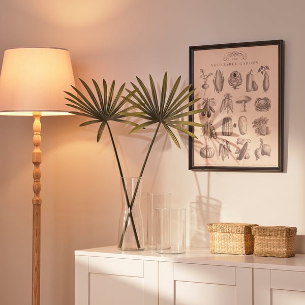 A cozy corner with a wooden floor lamp, vases with palm leaves, a vegetable garden poster, and wicker baskets on a sideboard.