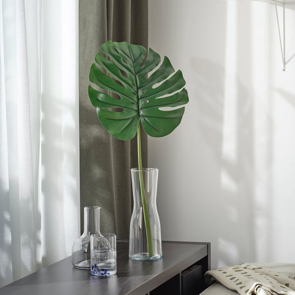 Large artificial green leaf in clear glass vase on dark shelf, with two smaller glass containers; modern decor.