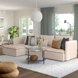 Beige SÖDERHAMN sectional sofa with colorful pillows, glass coffee table, woven ottoman, large windows with grey-green curtains.