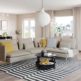 Modern living room with SÖDERHAMN sofa in beige, yellow & black pillows, striped rug, hanging lamp, large windows, plants, and coffee table.