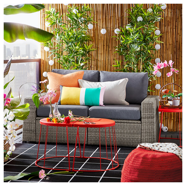 Modern outdoor living area with grey SOLLERÖN sofa, colorful pillows, red metal tables, lush plants, and city views.