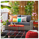 Modern outdoor living area with grey SOLLERÖN sofa, colorful pillows, red metal tables, lush plants, and city views.