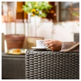A person holding a white coffee cup at an outdoor table with a yellow plate, brown wicker furniture.