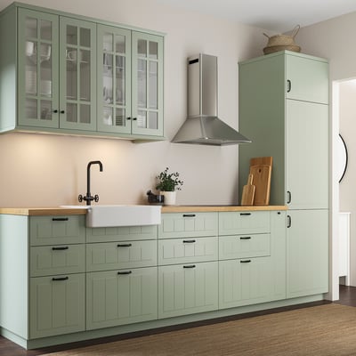 Modern kitchen with green cabinets, wood counter, white sink & black faucet. Tall units with glass doors and centered range hood.