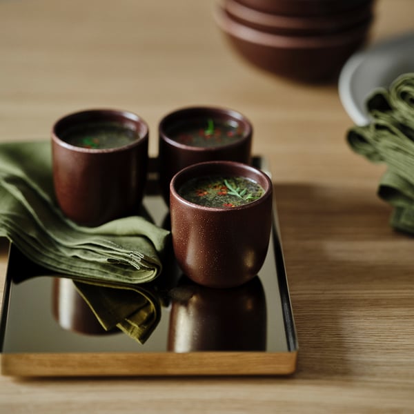 Three brown ceramic cups hold green herbs on a wooden tray, with a green linen napkin.