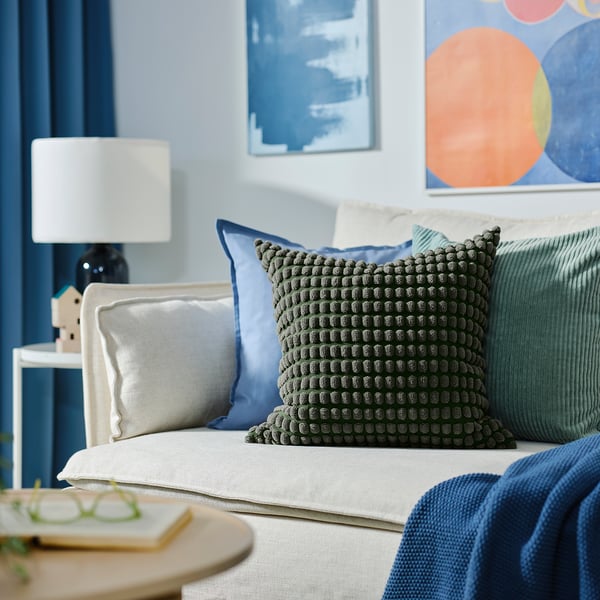 Bedside with cushions, lamp, and art. Grey-green pom-pom pillow, blue pillow, white lamp, colorful art.
