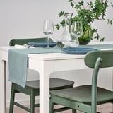 Table set with white table runner, blue plates, green glassware, and green chairs. A clear decanter and green vase with plants are also present.