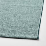 A close-up view of a textured, green-blue cotton table runner. The fabric appears soft, dense, and well-crafted with visible stitching.