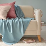 Light blue THORGUN fleece blanket draped on beige chair, designed for softness and warmth, made from recycled polyester.