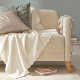 Blanket on light chair with pillow. Soft, white fleece, recycled polyester. Simple, comfortable design. A couple of notebooks lie beneath.