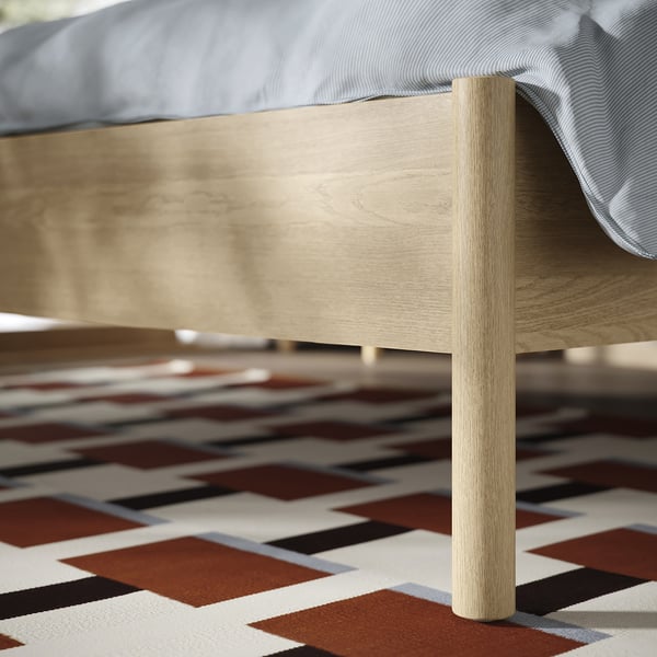 Modern, sturdy wooden bed with clean, light finish and visible wooden leg, on patterned rug.