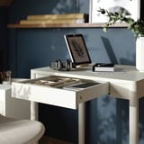White desk with open drawer containing pens, a tablet, and headphones.
