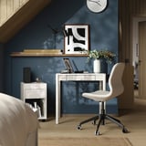 Modern home office with white TONSTAD desk, chair, and dark blue wall. Desk has a laptop, flowers, and a clock.