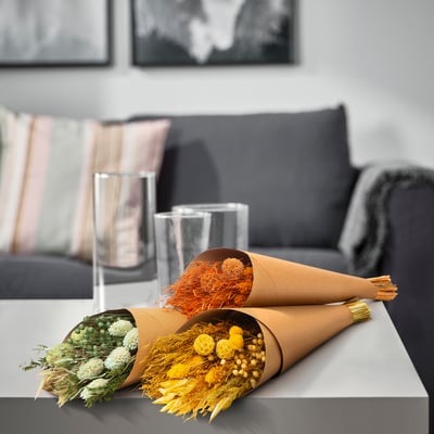 Two dried flower bouquets in brown paper cones on a gray table in a living room setting, with a black couch and pillows behind them.