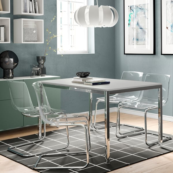 Modern dining set with clear chairs around white TORSBY table, chrome legs, gray rug, minimalist art.