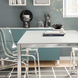 Modern metal table with clear chairs, small decorative items on top, grey wall behind.
