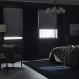 Modern bedroom with black walls and TREDANSEN blinds partially drawn, blocking light. A bed with a blanket and a chair are visible.