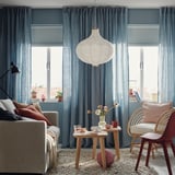 Modern room with TREDANSEN blue curtains, grey sofa, wooden table, wicker chair, rug, and hanging lamp. Blinked curtains frame large windows.