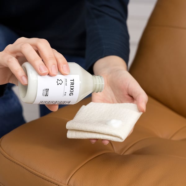 Person applies leather conditioner from a bottle labeled TRIXIG onto a cloth, preparing to clean and protect leather furniture.