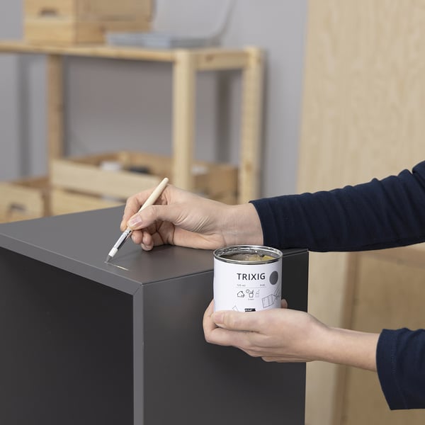 Person marking furniture with pen, holding TRIXIG paint can, in craft room.
