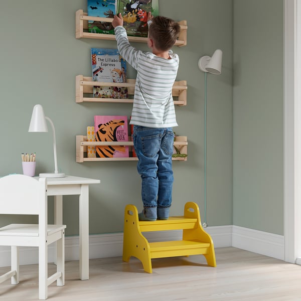 Child standing on yellow TROGEN stool, reaching for books on wall shelves in a green room.