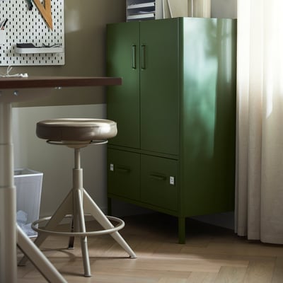 A modern green office setup includes a tall cabinet, a table with a swivel chair, and a pegboard storage system.