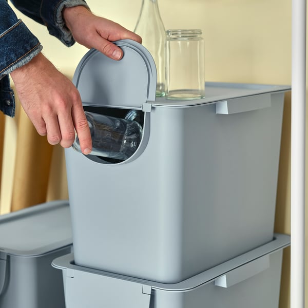 Person discarding glass bottle into gray TRÖSKNING recycling bin with lid open, showing waste sorting functionality.