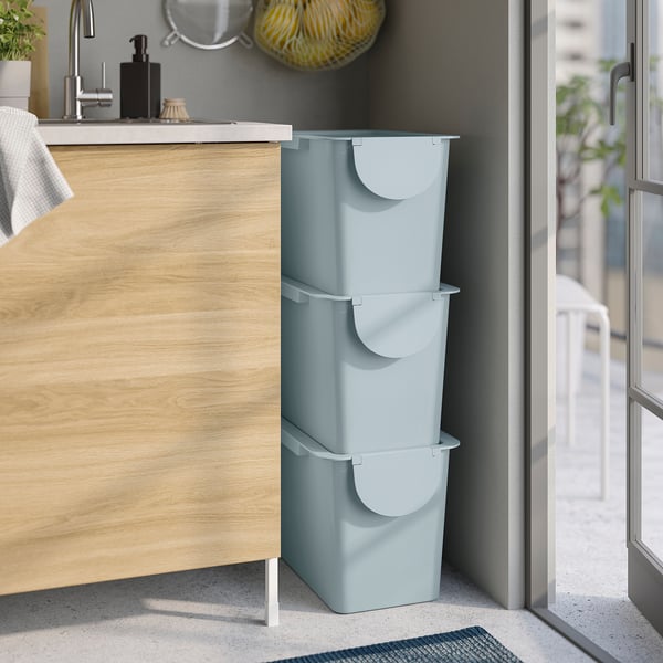 Three light blue, stackable bins with lids, suitable for waste sorting or storage, placed beside a wooden cabinet.