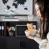 Woman eating noodles from a bowl while sitting in front of an office desk with a computer and bookshelf in the background.
