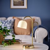 TVÅMASTAD lamp on table with couch & plant. Gold lamp with frosted glass shade and portable handle.