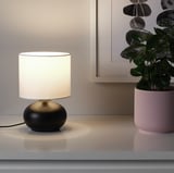 A small table lamp with a black stoneware base and a white lampshade, emitting a warm light.