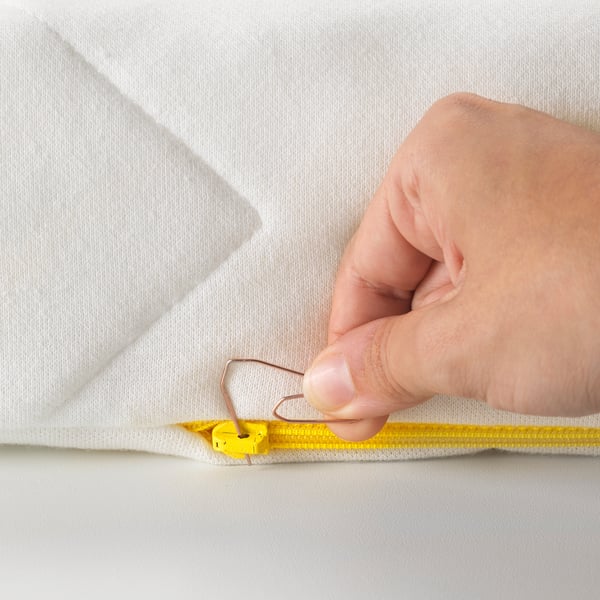 A hand is shown threading a paper clip to open a child-safe zipper on a white mattress cover.