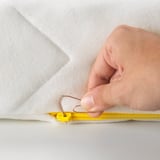 A hand is shown threading a paper clip to open a child-safe zipper on a white mattress cover.