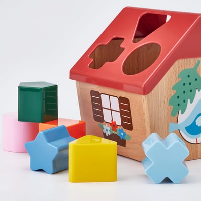 A red UPPSTÅ shape sorter toy resembling a birdhouse with shapes around. Its for learning shape and color recognition.