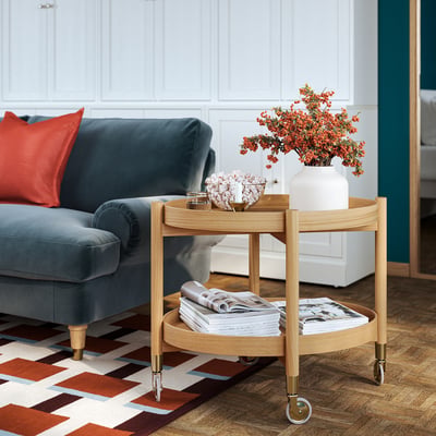 The room a VALNÄS blue sofa with a red pillow, a wooden coffee table with magazines and orange flowers, and a geometric rug.