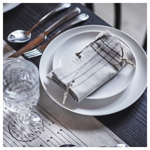 A table setting with VÅRARV beige and white napkin, on a white plate, alongside cutlery and a wine glass.