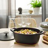 Black cast iron casserole with roasted veggies on wooden table, next to wine glasses, plates, and bread.