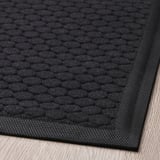 Dark, textured doormat with patterned surface and non-slip backing on wooden floor.