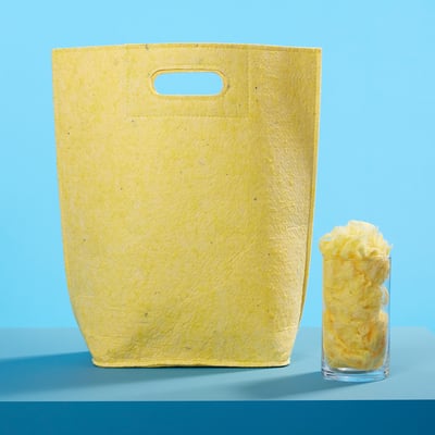 Large yellow VÄXELBRUK bag, made from recycled fibres, stands tall beside a smaller glass of yellow shreds on a blue surface.