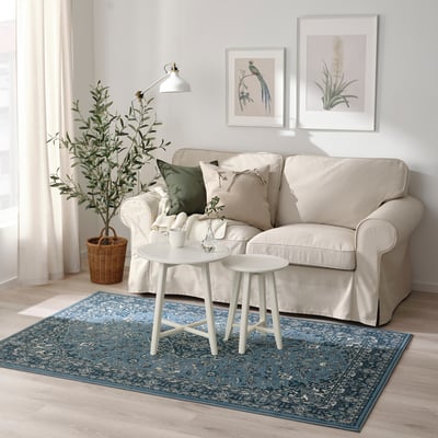 Cozy living room with beige sofa, white coffee table, blue floral VEDBÄK rug, art, plant.