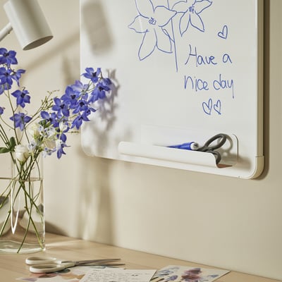 Small white VEMUND magnetic pen holder attaches to a board, holding a pen and scissors near a blue flower drawing message board, light grey wall.