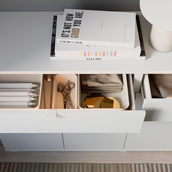 Modern white storage sideboard with drawers and open shelves. Top holds stylish books, drawer contains scissors, fabric, and circles.