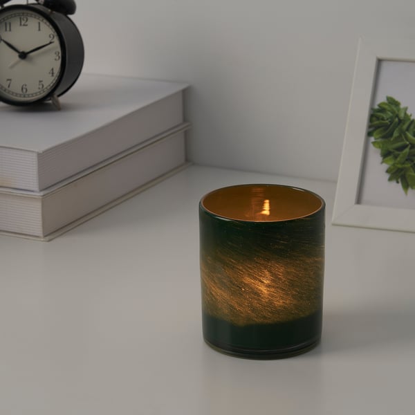 Lit candle in green glass holder beside books, clock, and framed plant. Provides warm, ambient light.